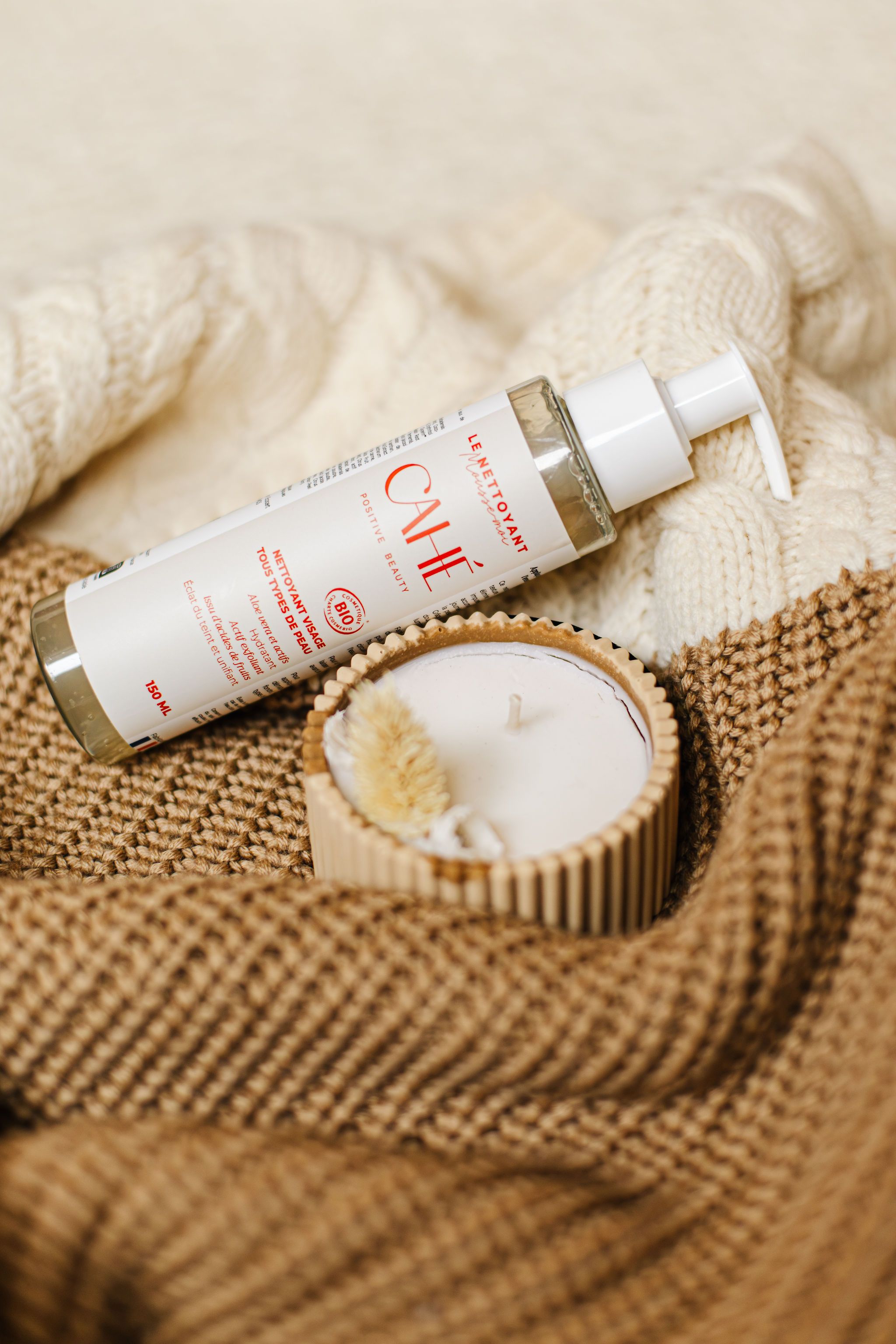Produit Cahé cosmetics disposé près d'une bougie sur un pull d'hiver aux tons blanc et vert