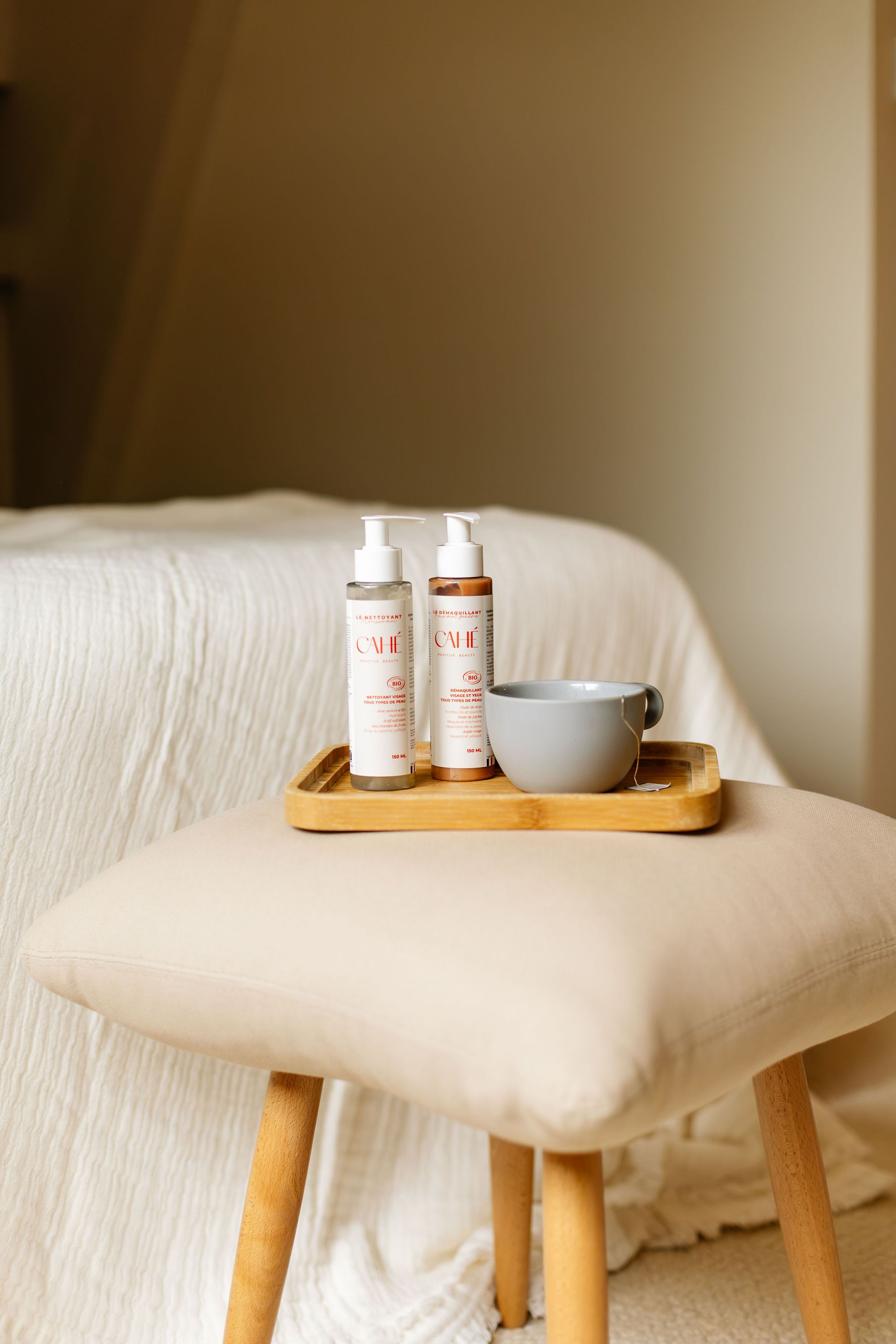 Produits Cahé Cosmetics disposés sur un plateau sur un petit pouf près d'un lit, dans une chambre aux tons épurés