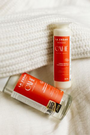 Crème de soin Cahé Cosmetics et sa recharge disposées sur un lit aux draps blancs, près d'un pull en laine blanc écru