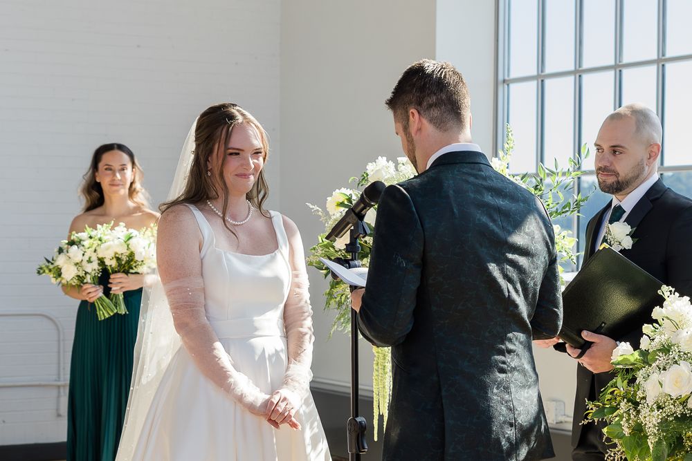 Smiling bride in a white lace gown holds hands with the groom during an outdoor wedding ceremony as a bridesmaid in green looks on.
