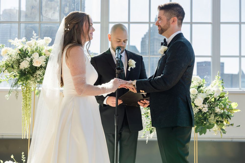 Bride and groom exchange vows while holding hands before an officiant at an elegant indoor wedding ceremony with white floral arrangements.