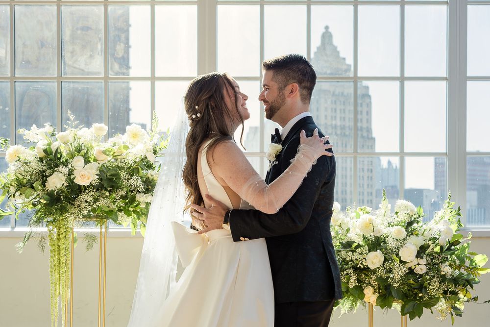 Bride in a white gown and groom in a black tuxedo embrace near large windows with white floral arrangements and a city skyline behind them.