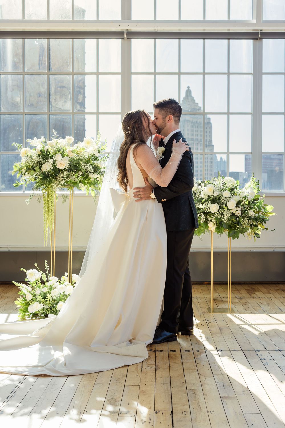Bride and groom kiss during their wedding ceremony in a bright loft venue with floral arrangements and city skyline views.