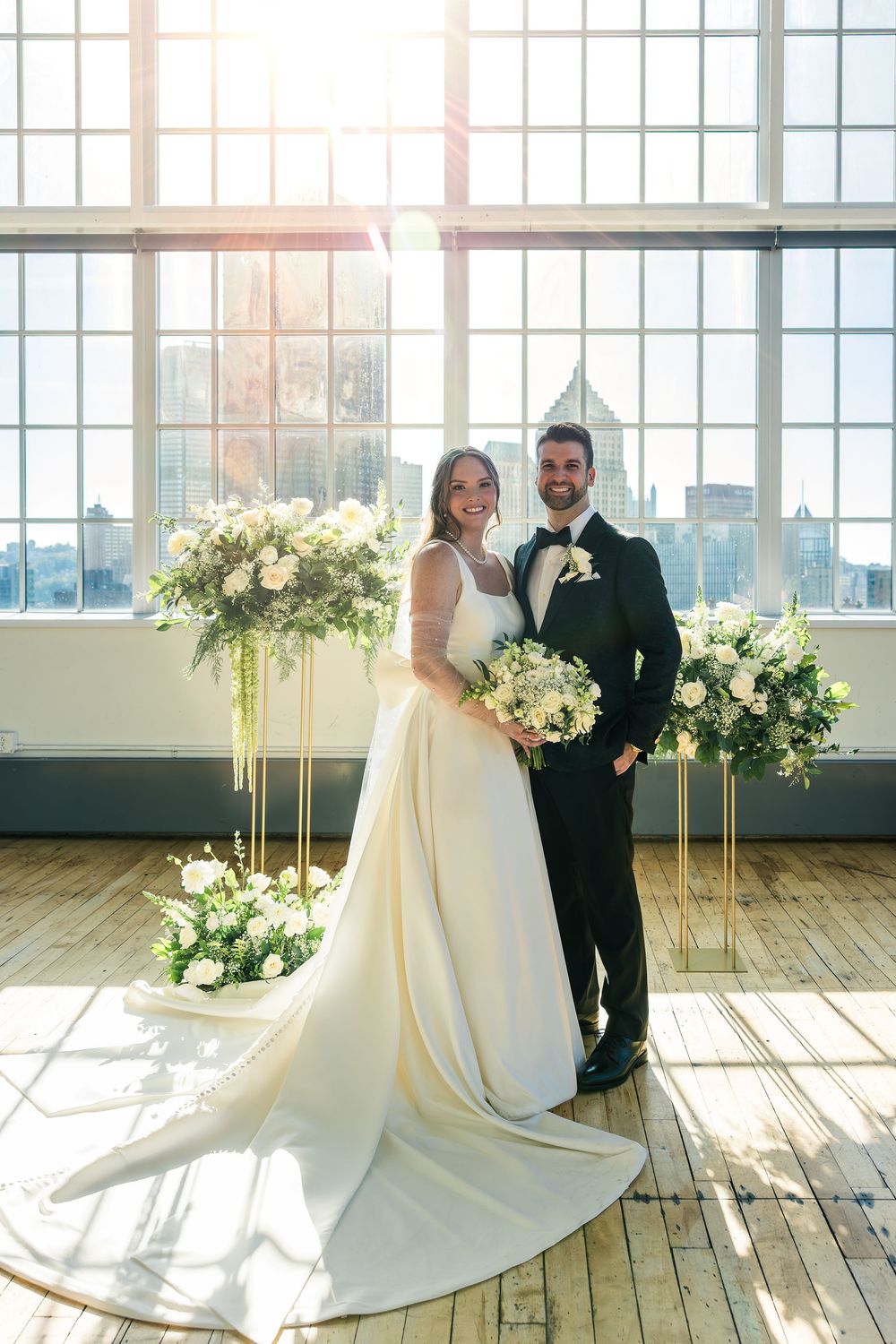 Bride in a white satin gown and groom in a black suit hold bouquets in a sunlit loft with floral stands and city skyline views.