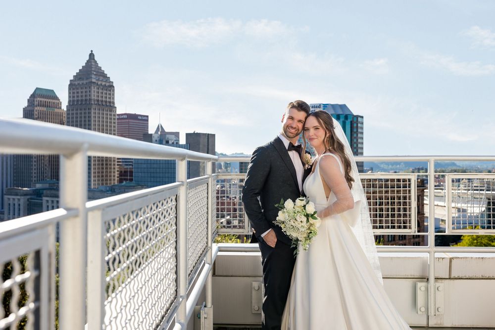 Bride in a white gown holding a bouquet stands beside her groom in a dark suit on a rooftop terrace with a city skyline behind them.