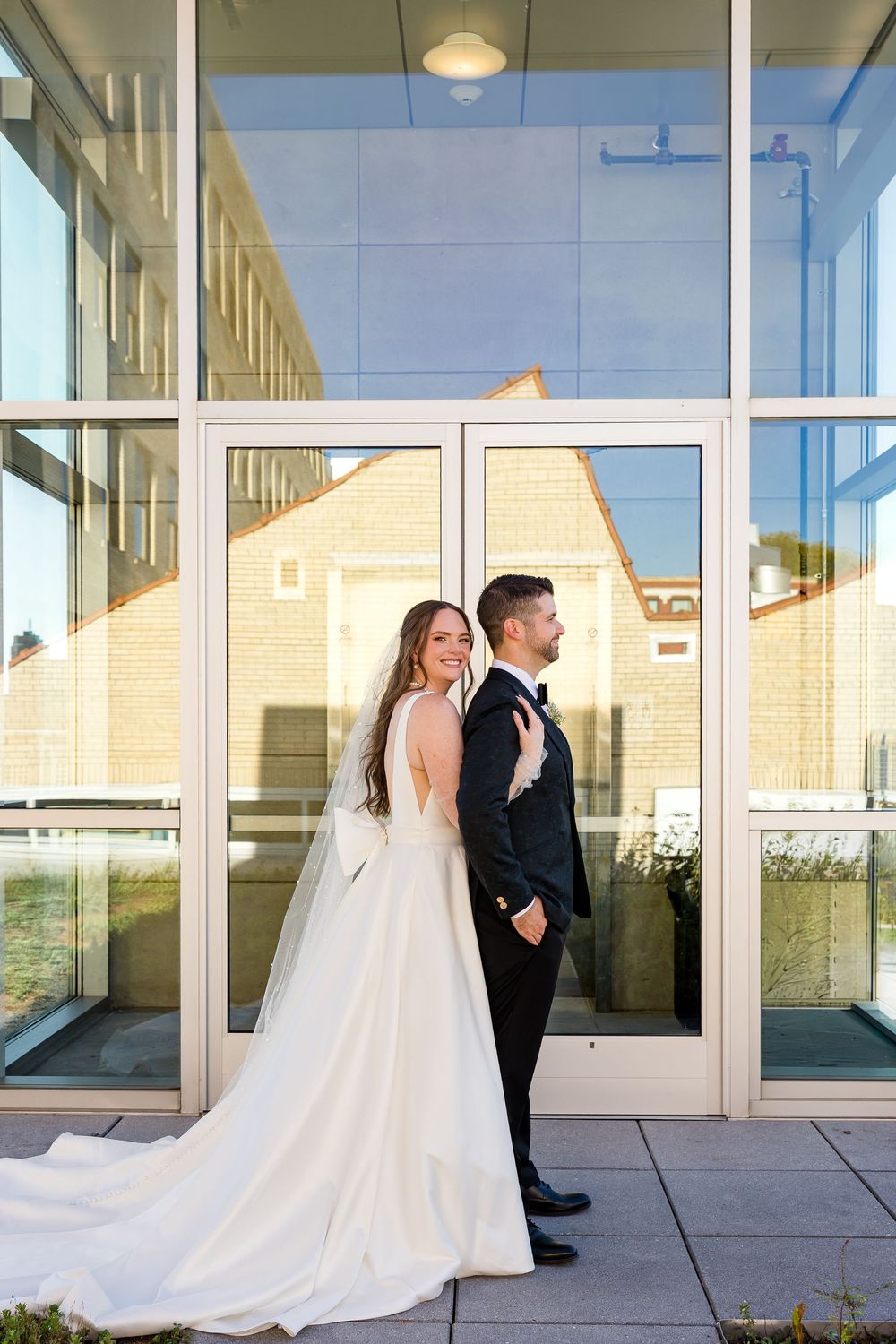 Smiling bride in a flowing white gown and veil poses with her groom in a black tuxedo in front of a glass building.