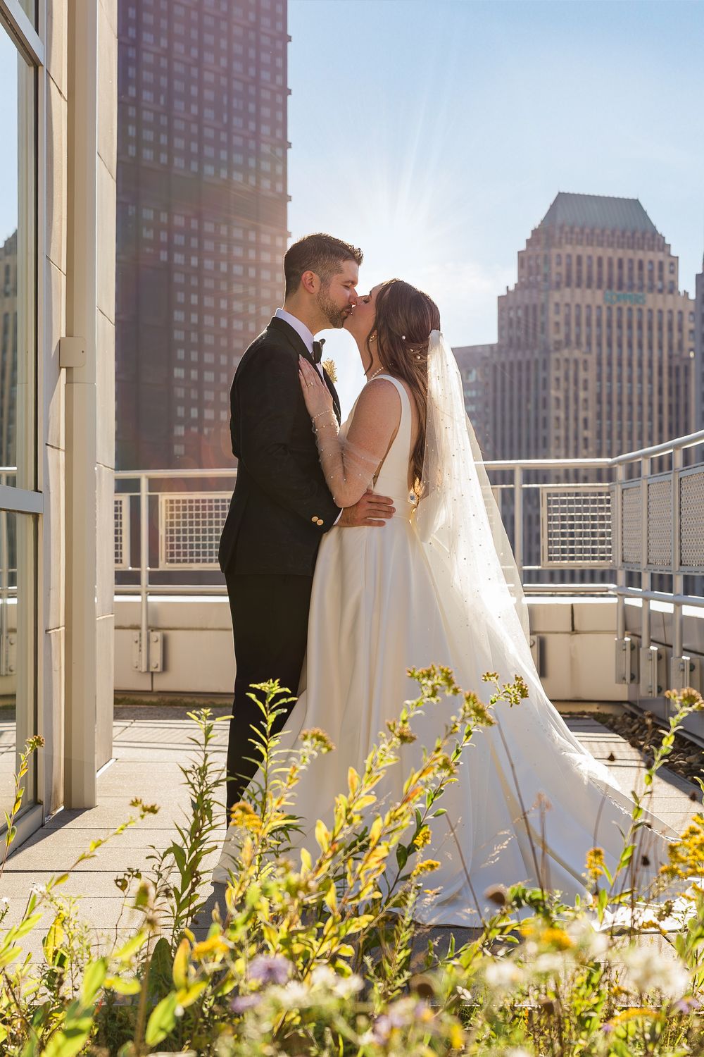 Bride in a flowing white gown and veil kisses her groom on a sunny rooftop terrace with city skyline views and wildflowers.
