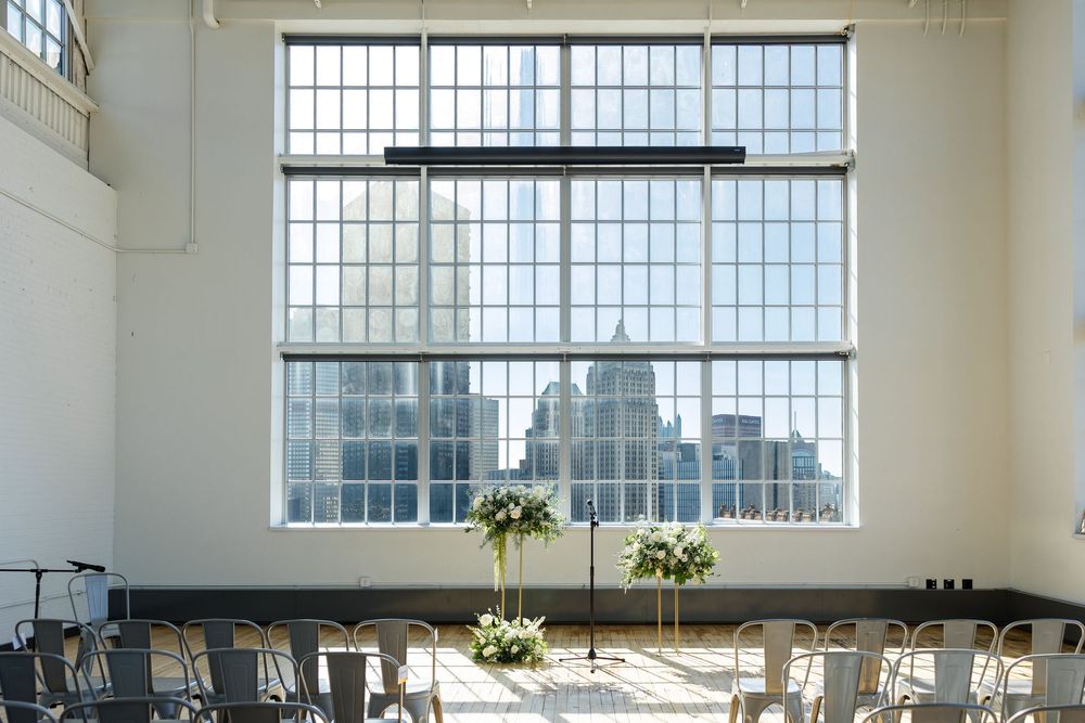 Bright industrial event venue with a large paned window overlooking the city skyline, floral arrangements, and chairs set up for a ceremony.