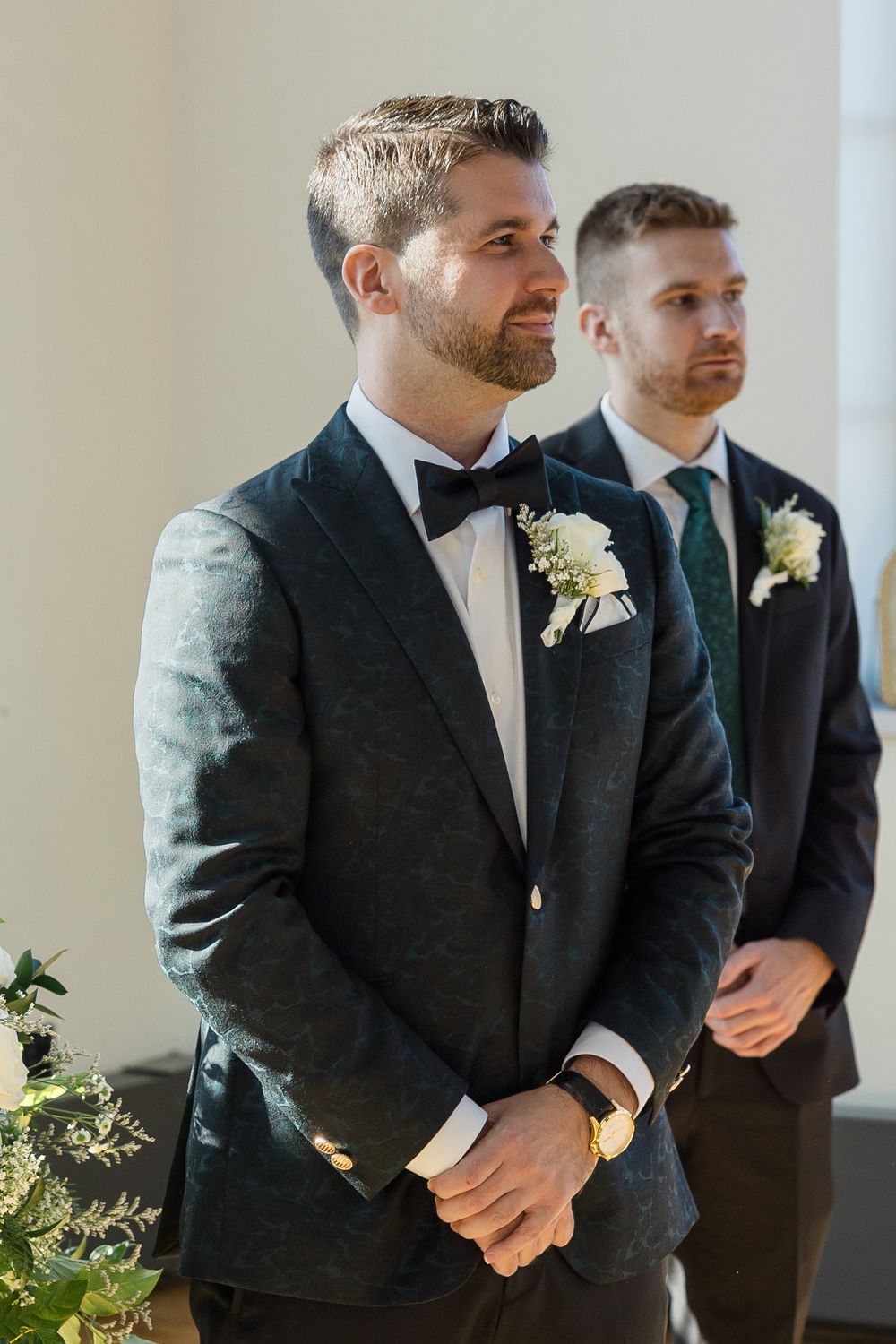 Groom in a dark green jacket, bow tie and white boutonniere stands at a wedding ceremony with a groomsman beside him.