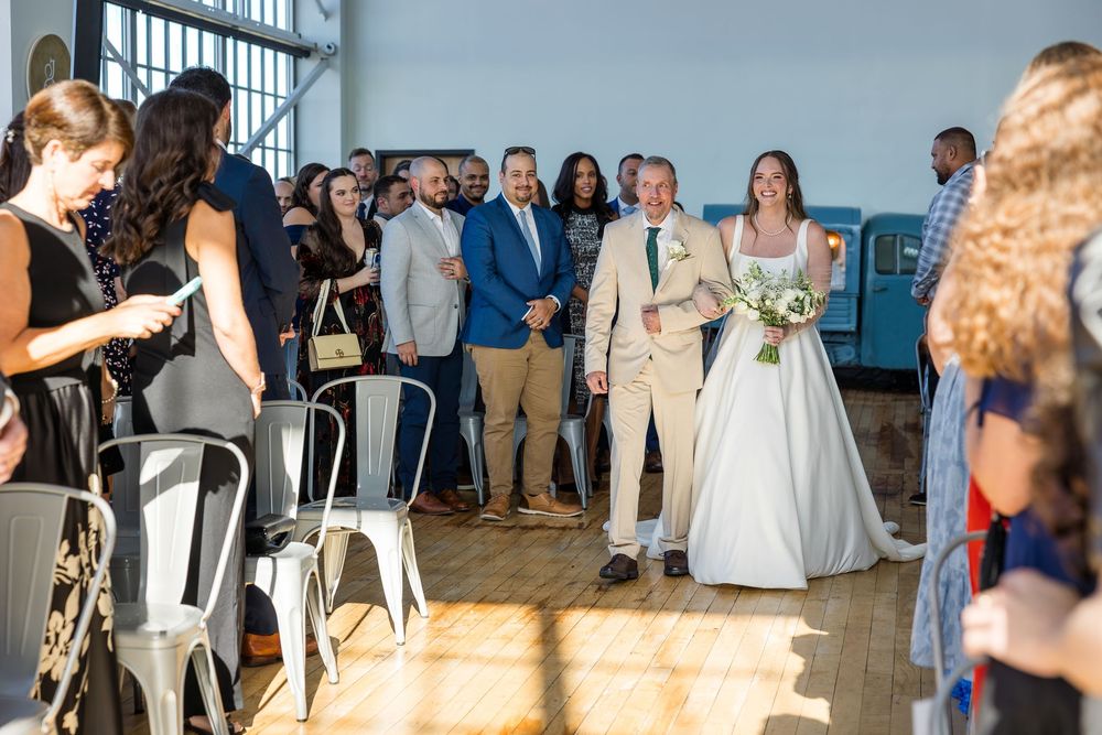 Bride in a white gown holding a bouquet walks down the aisle with an older man in a tan suit as wedding guests look on.