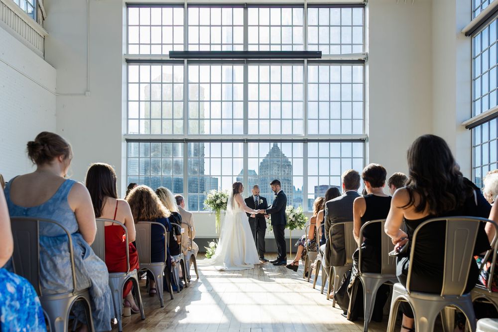 Bride in white gown and groom exchange vows before seated guests in a bright wedding venue with tall industrial windows.