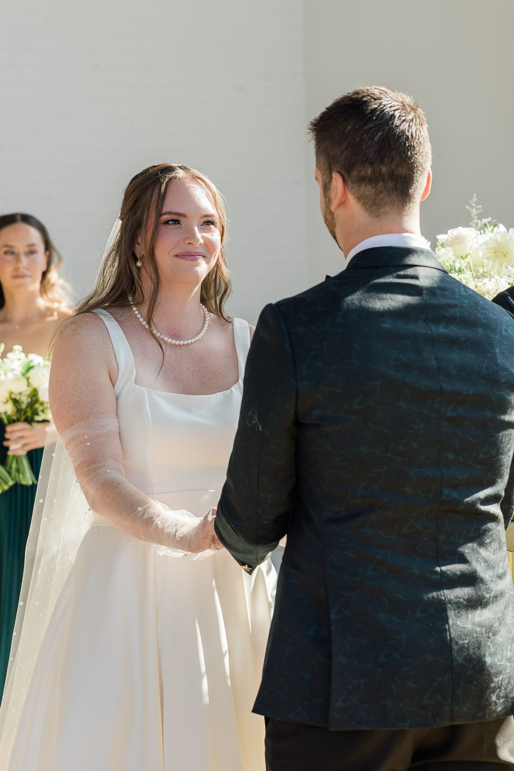 Smiling bride in a white satin gown and pearl necklace holds hands with the groom in a dark jacket during an outdoor wedding ceremony.
