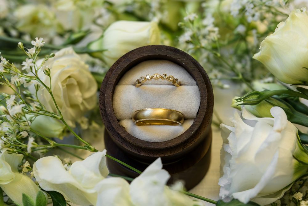 Gold wedding bands resting in a round wooden ring box surrounded by white roses and delicate baby's breath flowers.
