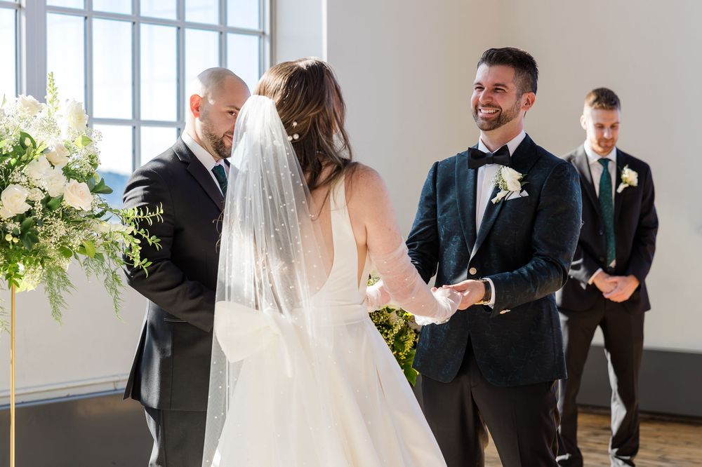 Smiling groom in a navy suit holds hands with a veiled bride in a white gown during an indoor wedding ceremony with floral decor.