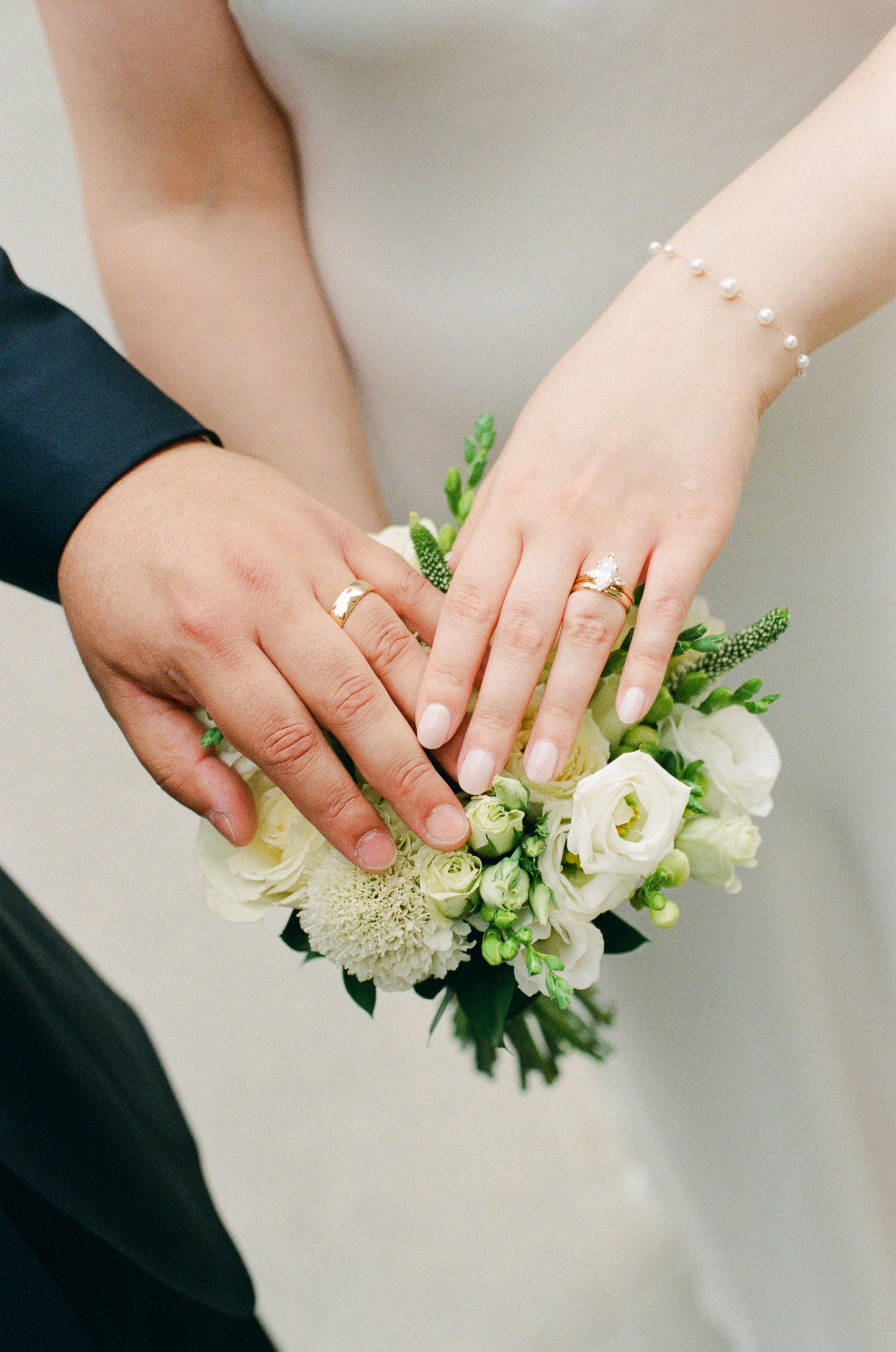 Newlywed couple displaying gold wedding rings on hands resting over a white bridal bouquet with green accents.