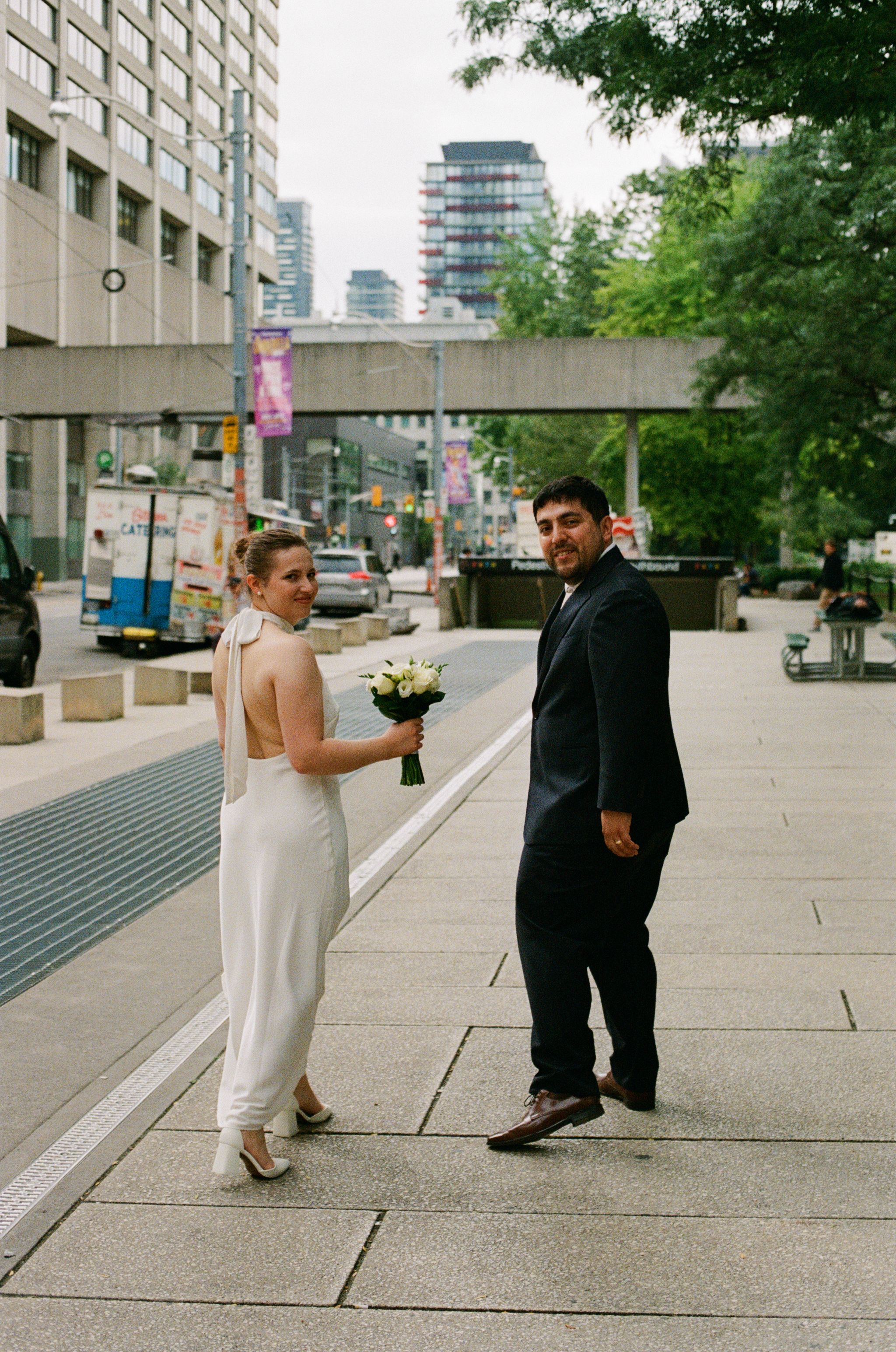 Bride in white backless dress holding bouquet and groom in black suit posing on urban plaza on wedding day.