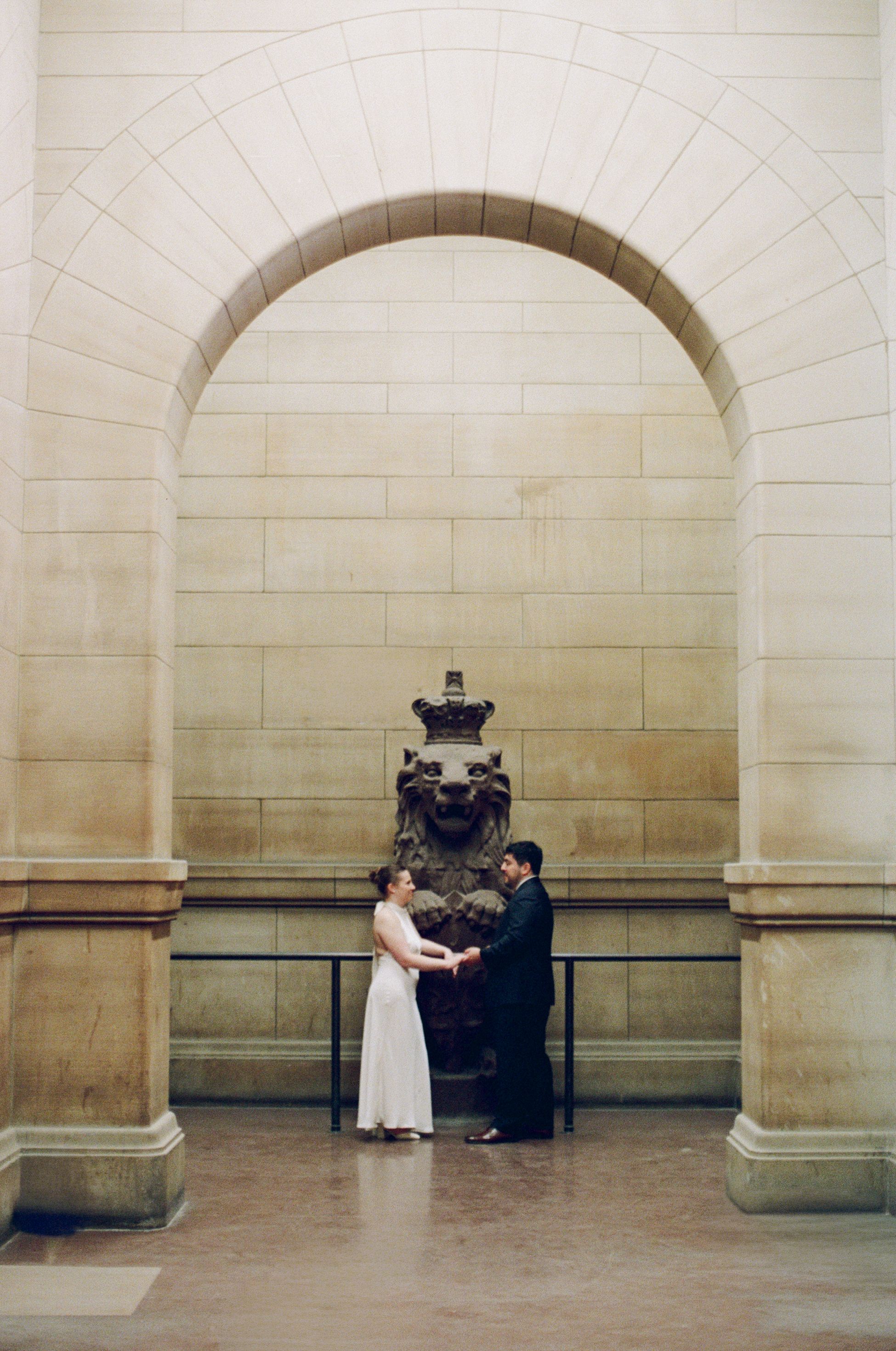 Couple holds hands before a bronze lion statue under an elegant stone arch in a grand neoclassical building.