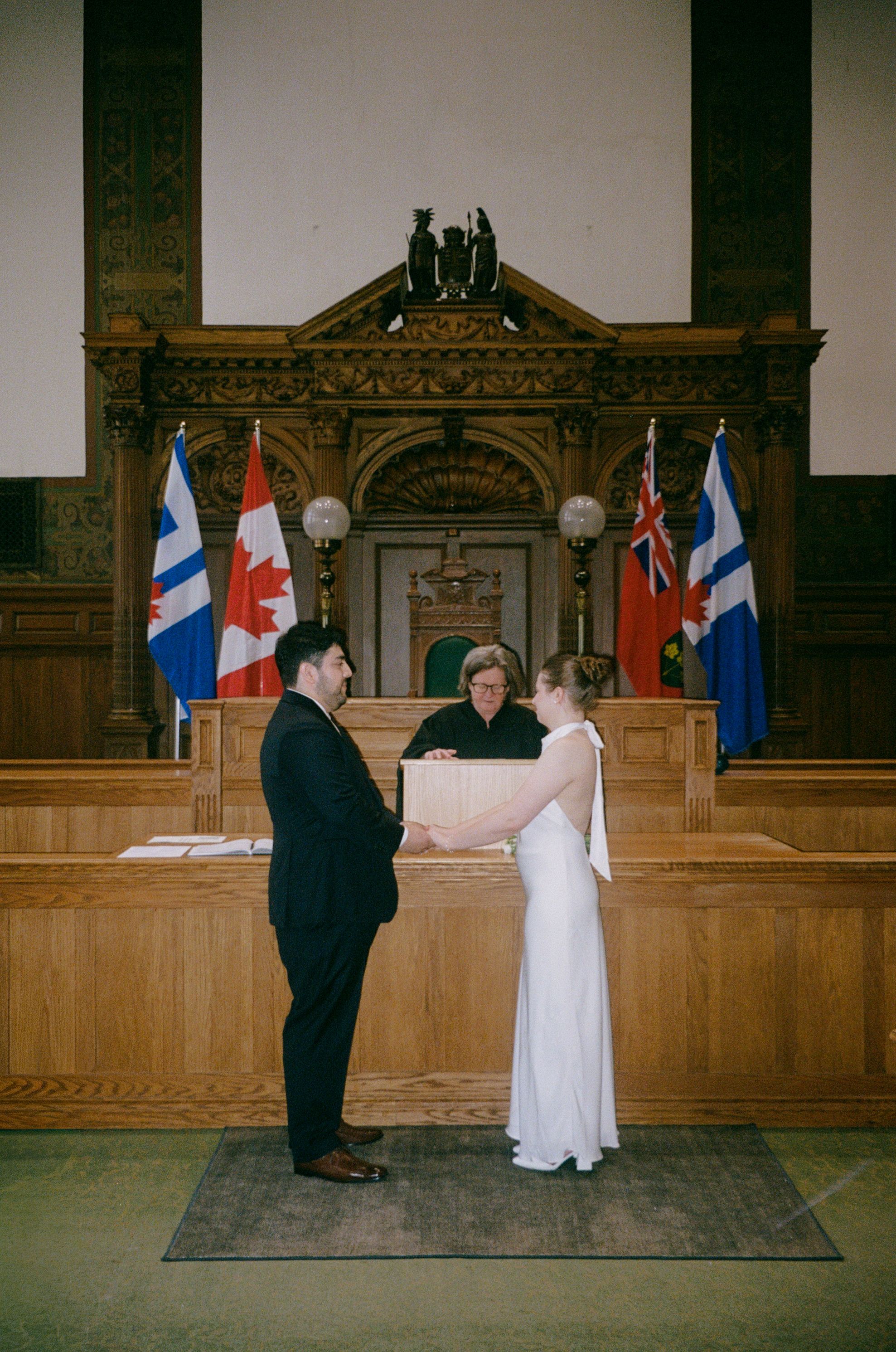 Couple exchanging vows before a judge in a wood-paneled courtroom with Canadian flags.