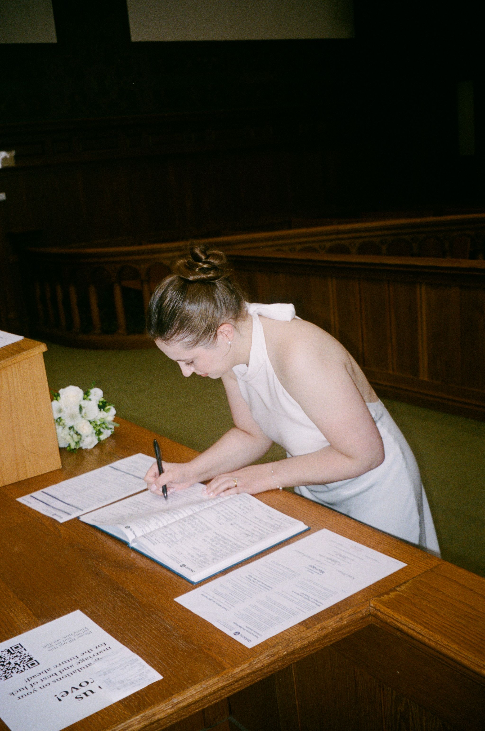 Bride in white dress signing marriage certificate at wooden desk with white flowers bouquet nearby.