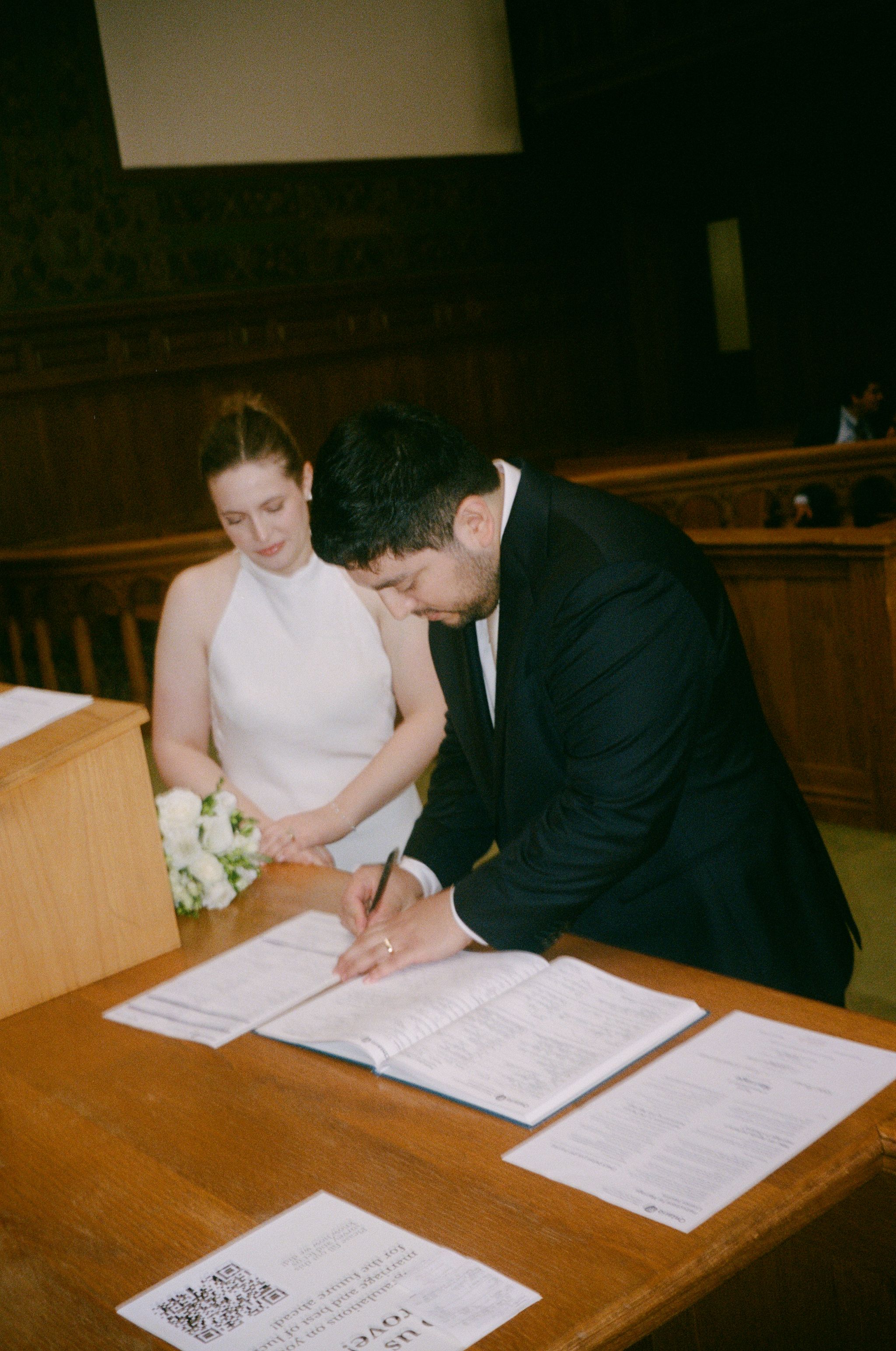 Bride and groom signing marriage certificate at wooden table during intimate courthouse wedding ceremony.