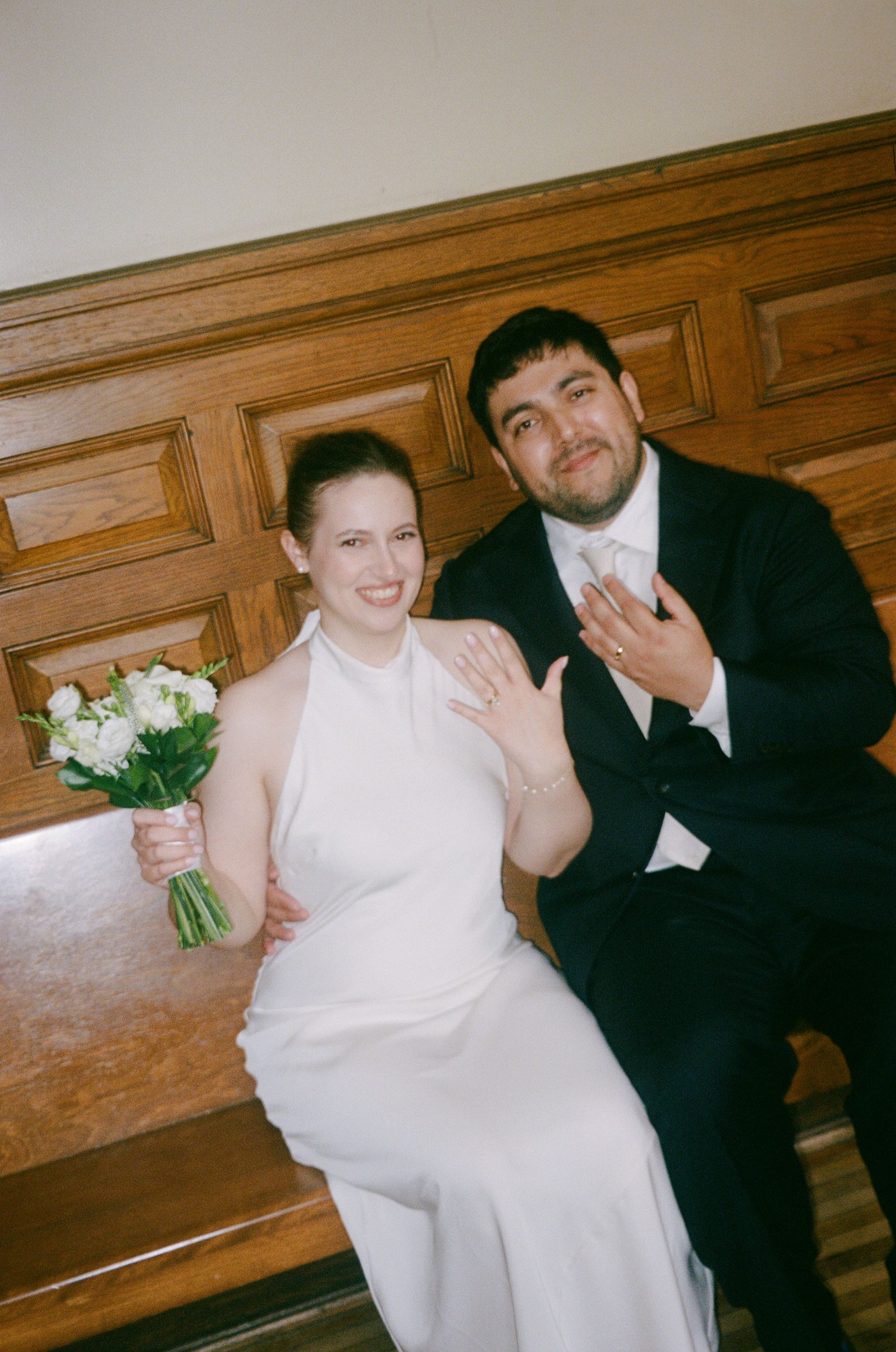 Newlywed couple sitting on wooden bench, bride in white halter gown holding bouquet, groom in black suit showing wedding rings.