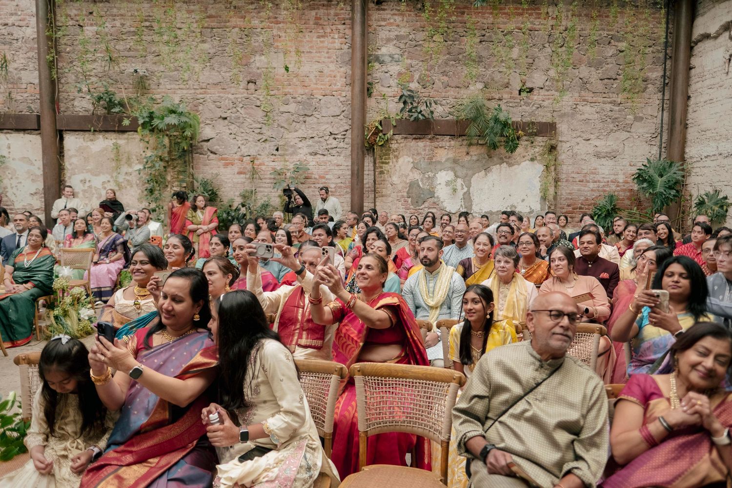 Large crowd of guests in colorful traditional Indian attire seated at an elegant wedding ceremony indoors.