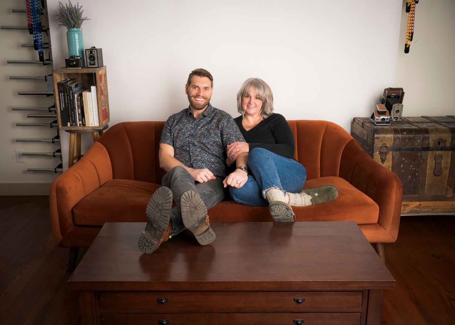 Smiling man and woman sit close together on an orange mid-century modern sofa behind a wooden coffee table in a cozy living room.