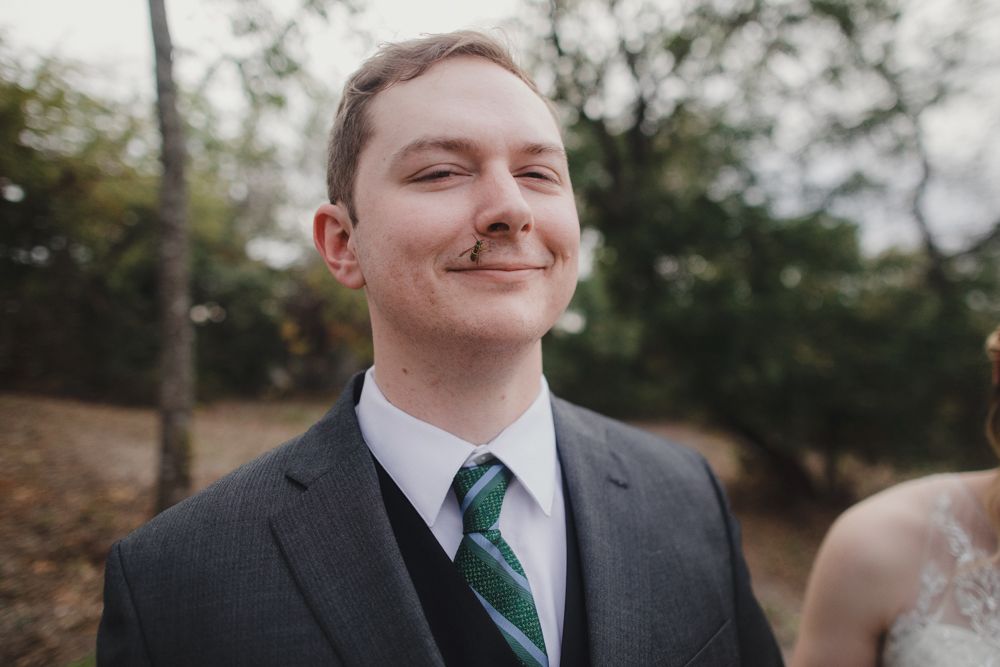 Groom in grey suit and striped tie smiling outdoors during autumn wedding.