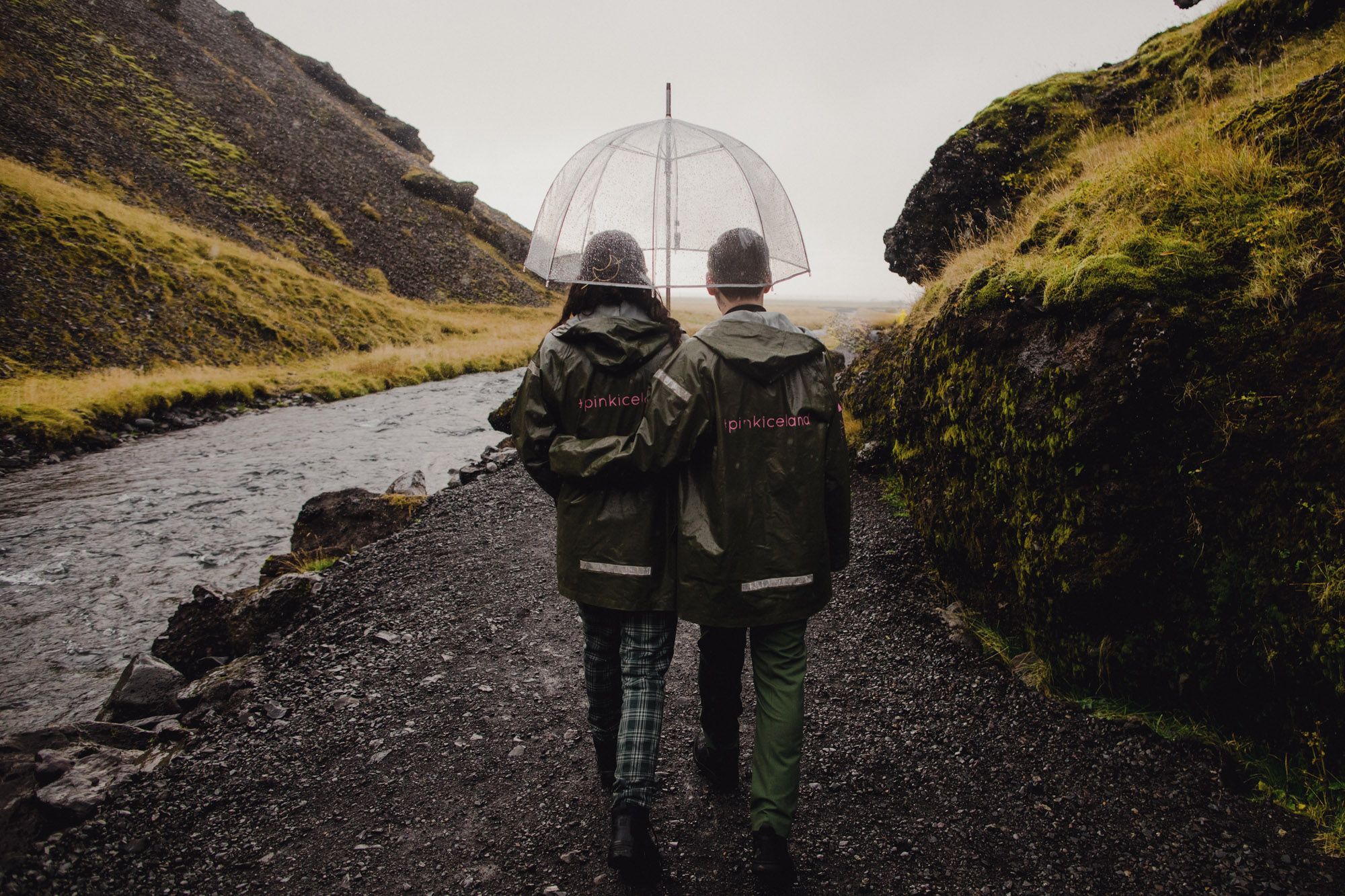 Two figures in matching raincoats walk along a scenic mountain path with clear umbrellas.