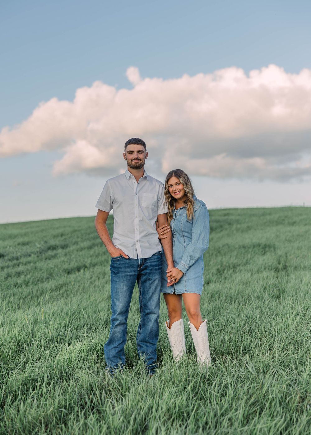 Smiling couple standing close together in a green grassy field under a blue sky with fluffy clouds, she wears a denim dress and white boots.