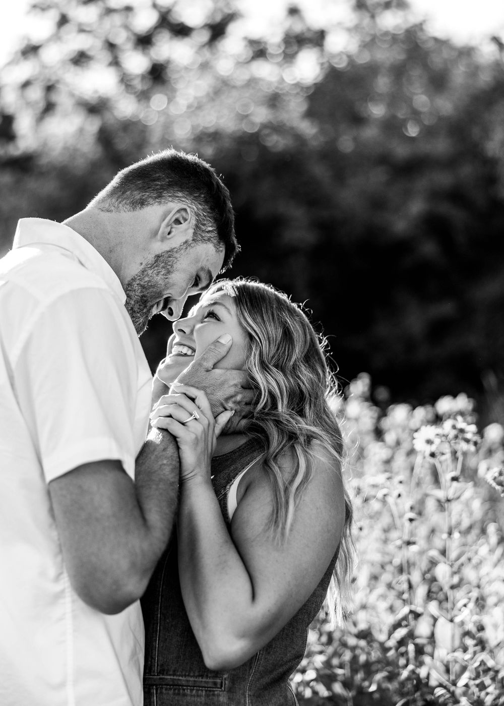 Black and white photo of a couple embracing outdoors, foreheads touching as she smiles, with soft blurred wildflowers in the background.