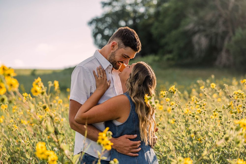 Romantic couple embracing and about to kiss in a golden field of yellow wildflowers during a warm, sunny outdoor engagement photo session.