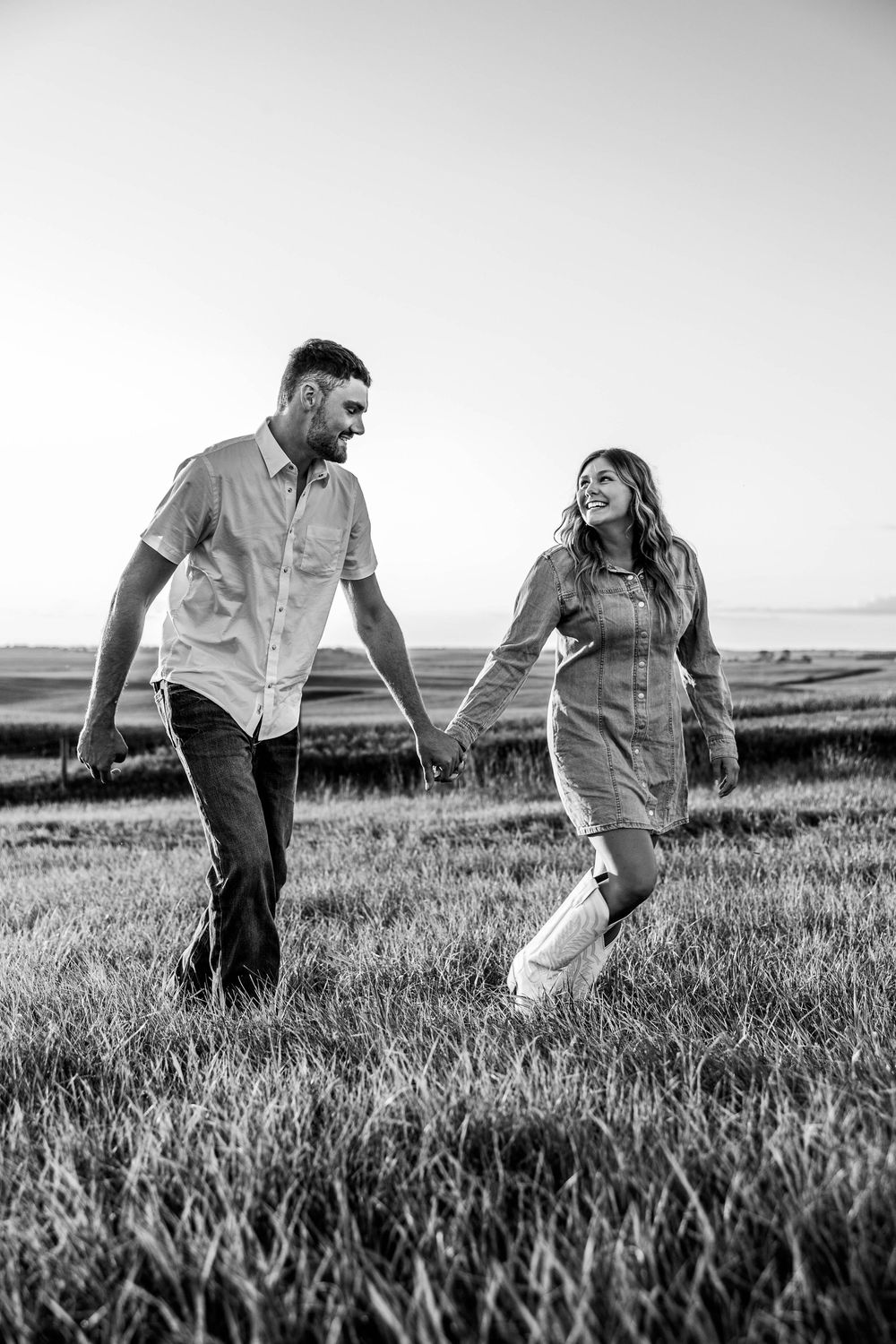Black and white photo of a smiling couple holding hands and walking through a grassy field at sunset during an engagement session.