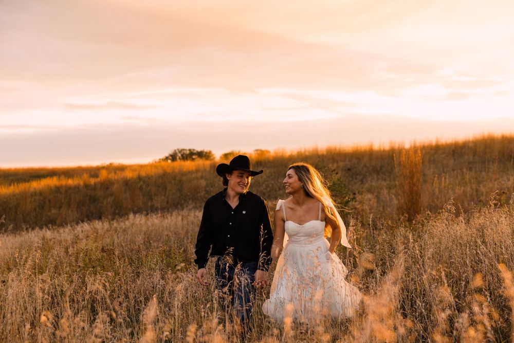Couple in a black cowboy hat and flowing white dress walk hand in hand through golden prairie grass at sunset.
