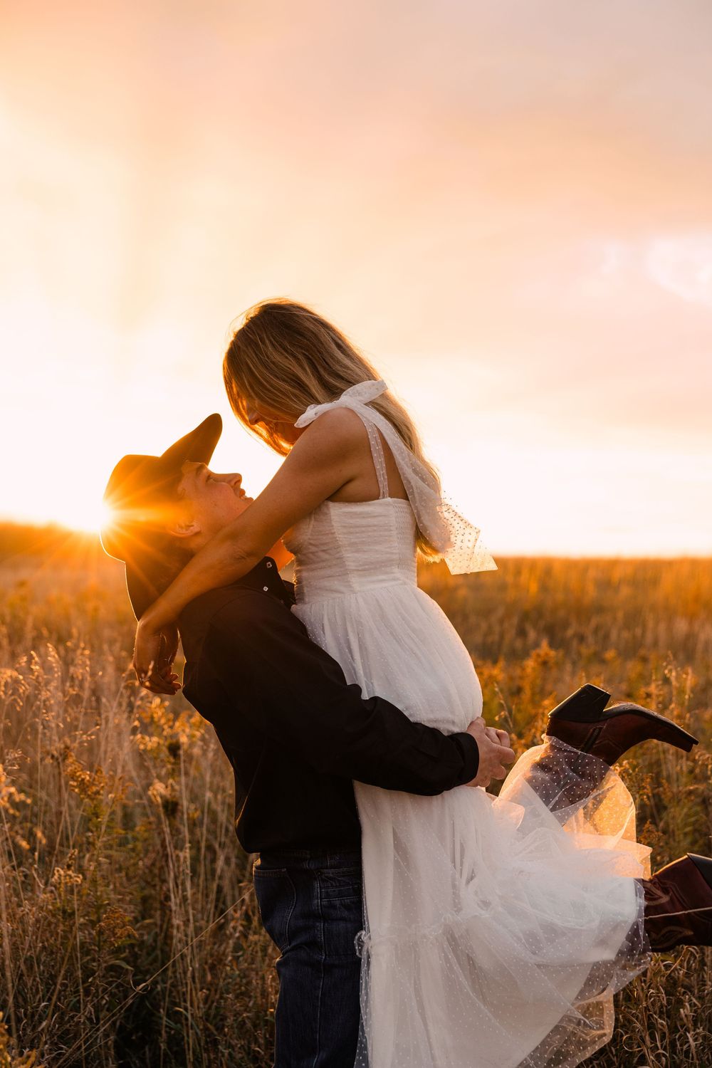 Man in a cowboy hat lifts a smiling woman in a white tulle dress in a golden field at sunset.