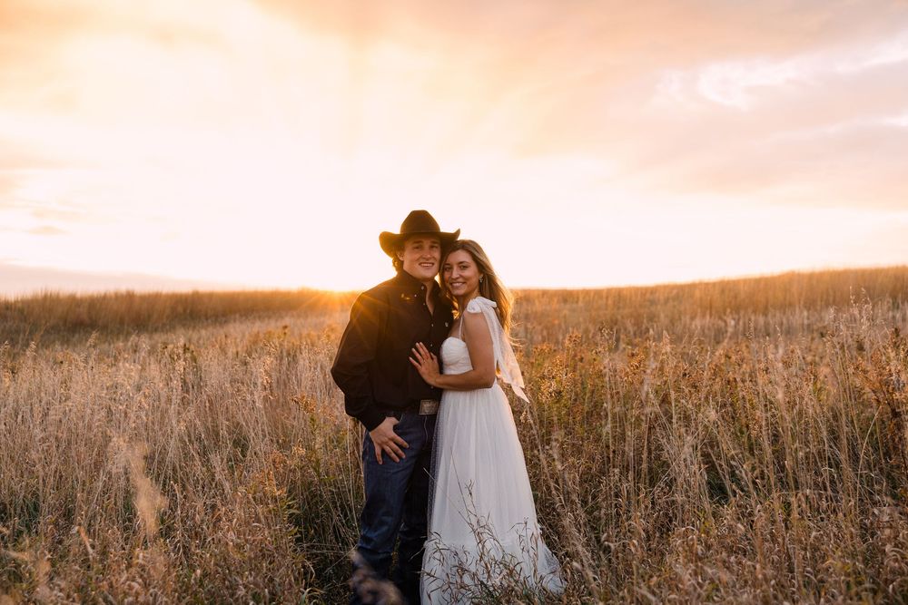Smiling couple embracing in a golden field at sunset, the man in a cowboy hat and dark jacket, the woman in a white dress.