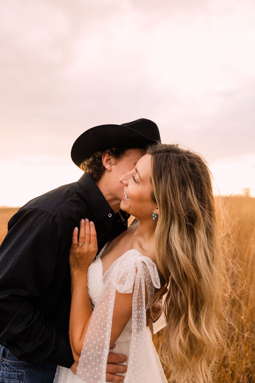 A smiling bride in a white dotted-tulle gown is kissed on the cheek by a groom in a black cowboy hat in a golden field at sunset.