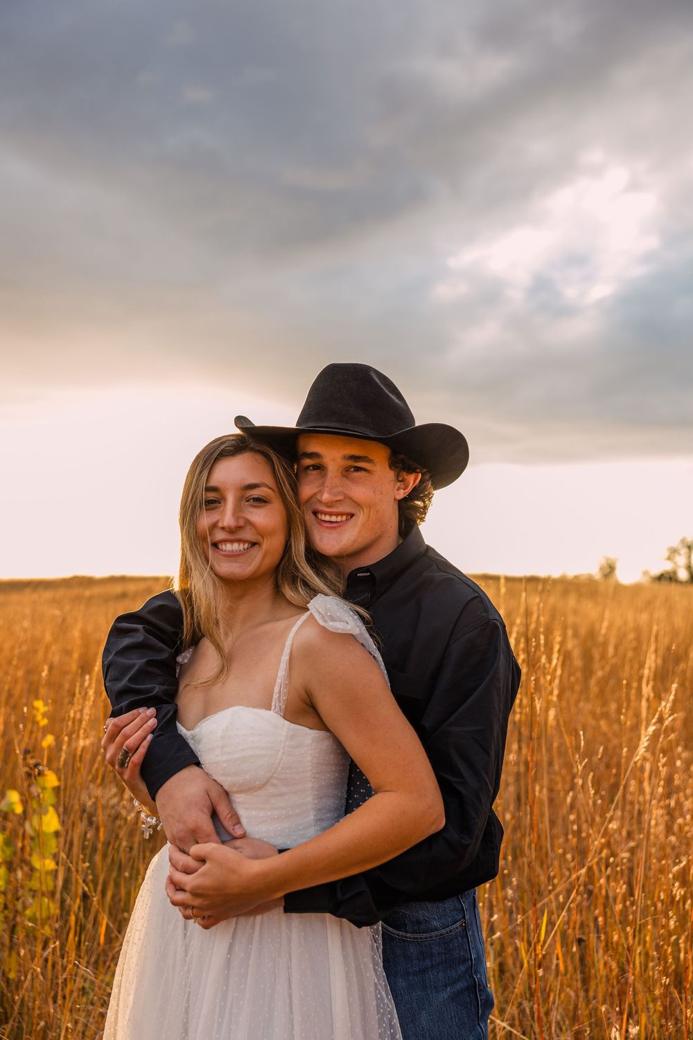 Smiling couple embracing in a golden wheat field at sunset, he wears a black cowboy hat and she a white dress, under a dramatic cloudy sky.