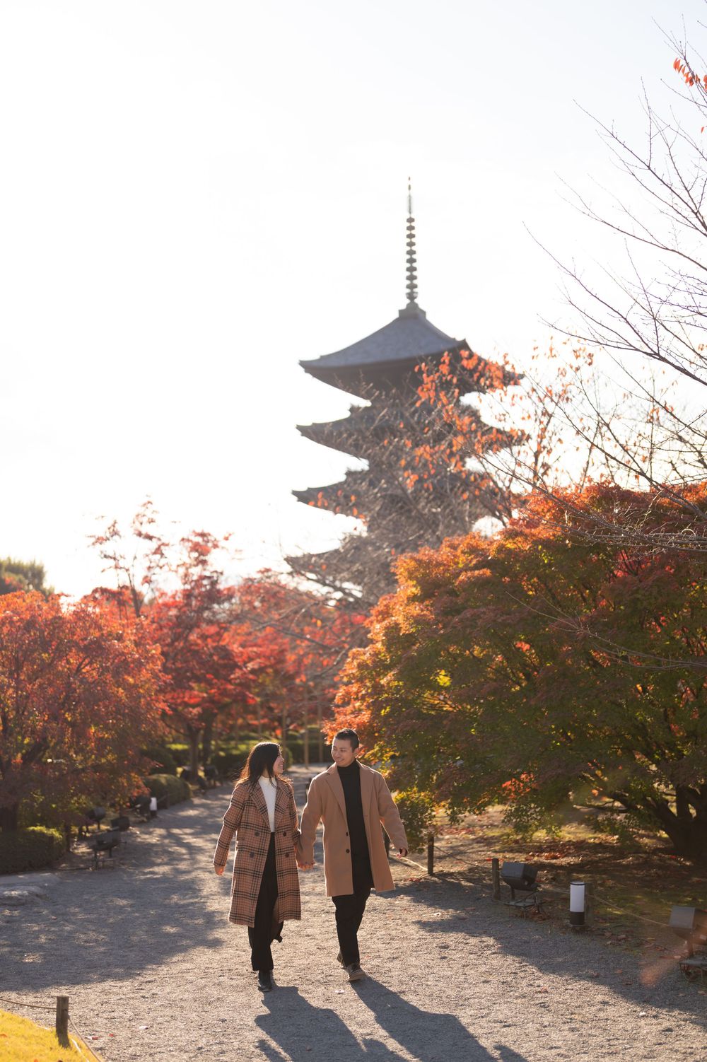 Couple in matching tan coats strolling a gravel path beneath vibrant red autumn maples with a tall Japanese pagoda in the background.