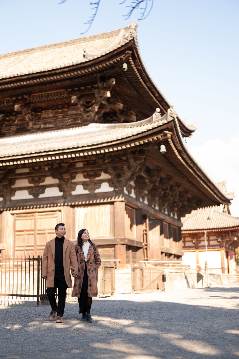 Couple in matching brown coats strolling past a traditional Japanese temple with ornate wooden eaves on a sunny day.
