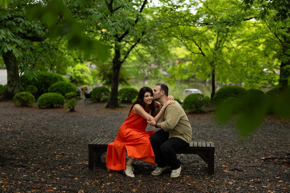 A couple in an orange dress and olive sweater share an intimate moment on a wooden park bench surrounded by lush green trees and shrubs.