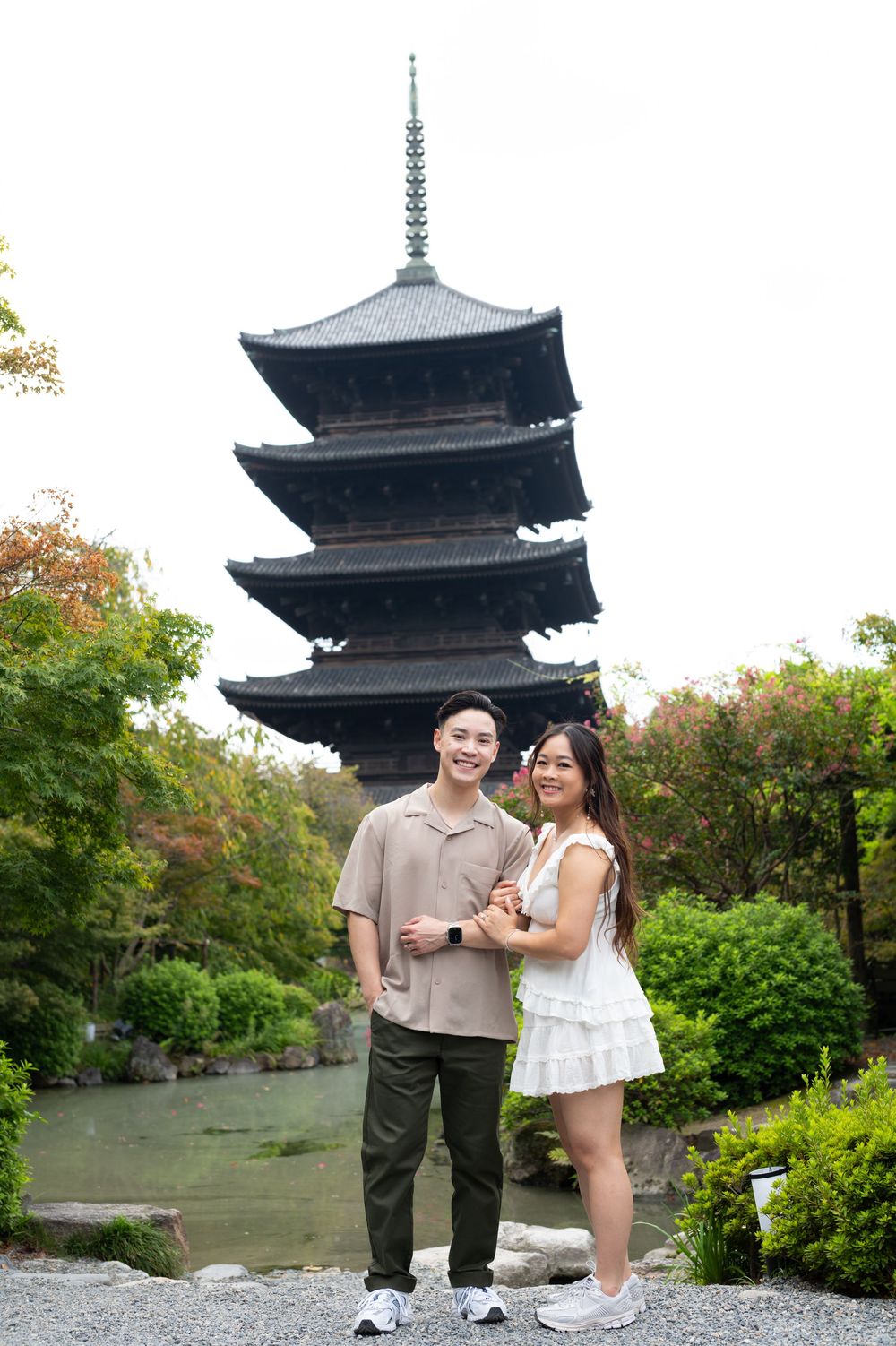 Smiling couple poses arm in arm near a pond with a traditional five-story Japanese pagoda rising behind lush green trees.