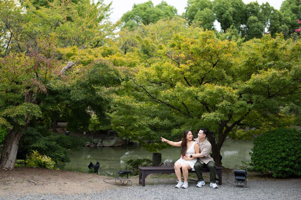 Couple sitting close together on a wooden bench in a lush Japanese garden, with a calm pond and vibrant green maple trees behind them.
