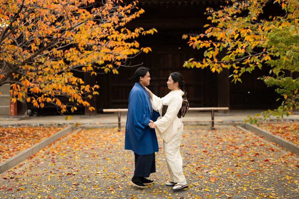 Couple in traditional Japanese clothing holding hands beneath vibrant orange autumn trees on a leaf-covered temple courtyard path.
