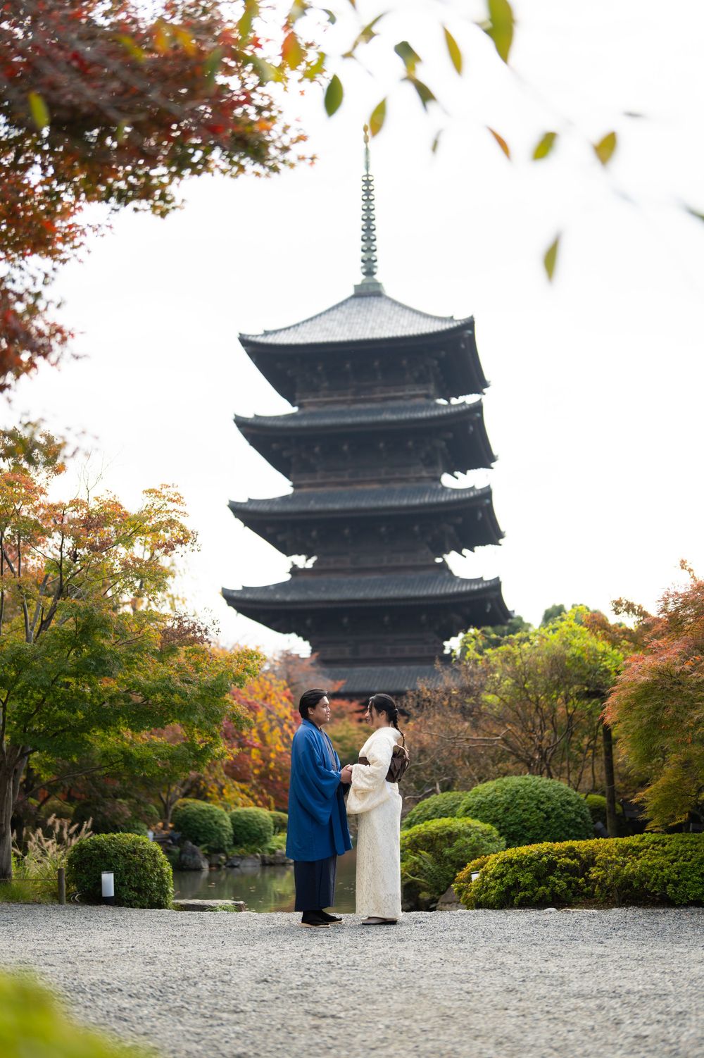 Couple in traditional blue and white kimono stand before a five-story Japanese pagoda surrounded by colorful autumn foliage.