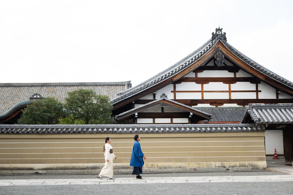 Two people in traditional Japanese clothing walk past a historic temple with a tiled roof and long earthen wall under a soft overcast sky.