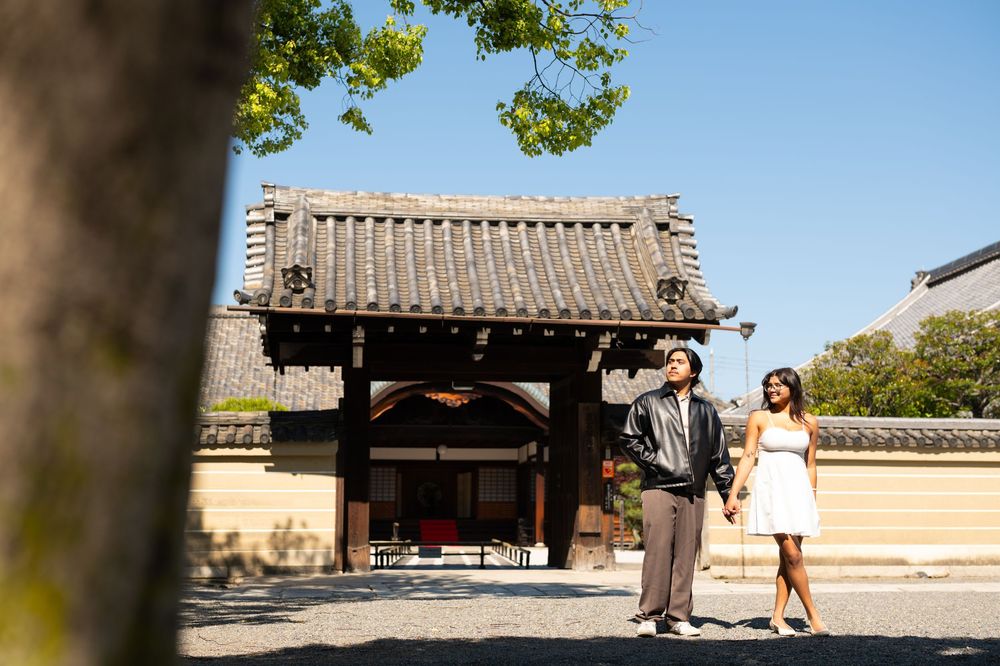 Couple holding hands and walking in front of a traditional Japanese temple gate with a tiled roof under a clear blue sky.