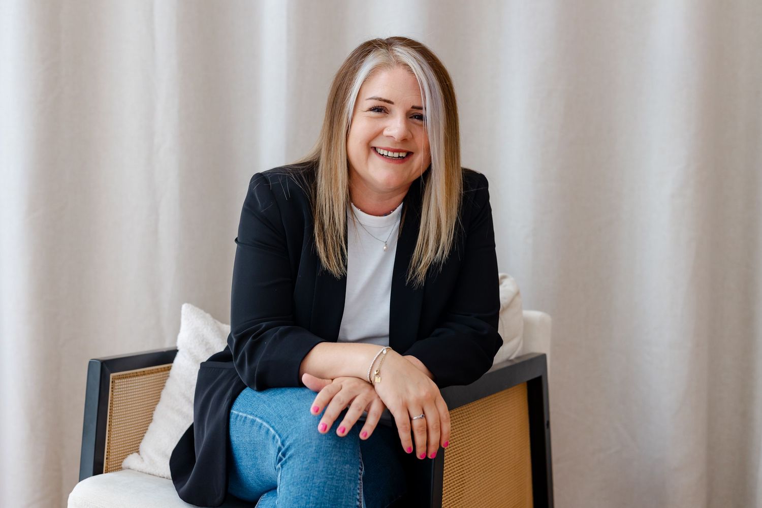 Smiling blonde woman in black blazer and jeans sits relaxed in white chair against neutral curtain backdrop.