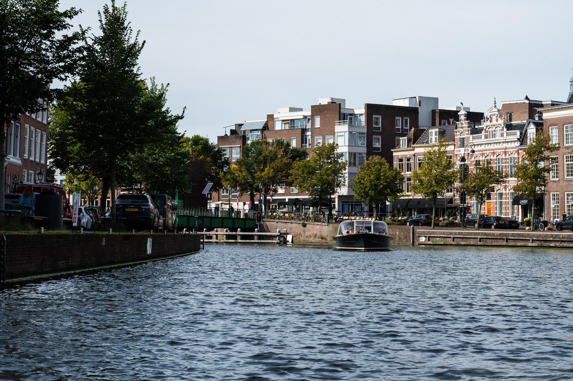 A scenic canal in Haarlem, Netherlands, with moored boats, tree-lined quays, and historic Dutch buildings under a cloudy sky.