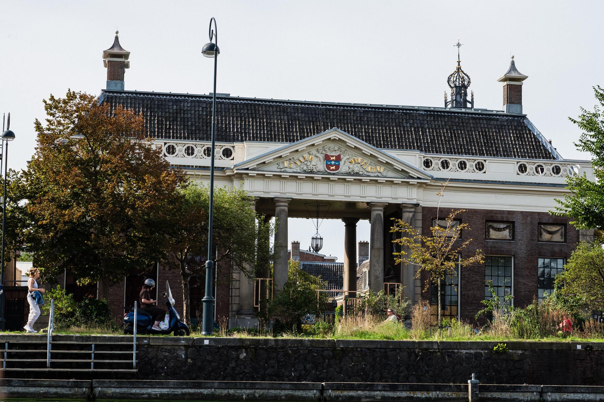Historic neoclassical building with columns and ornate roofline beside a waterway, with people relaxing on a sunny day.
